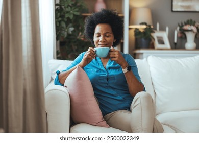 African Woman sitting on her sofa in her living room holding a cup. She relaxing. Portrait of a happy African American woman at home warming up her hands with a cup of coffee - Powered by Shutterstock