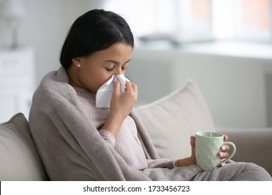 African woman sitting on sofa in living room wrapped in warm plaid hold paper handkerchief blow runny nose relief nasal stuffiness drinks cold medicine. Influenza flu symptoms, seasonal grippe concept - Powered by Shutterstock