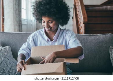 African woman sitting on sofa in living room hold on lap big carton box unpacking parcel received from online shopping. Curious black lady unpacking cardboard parcel. Smiling female unboxing package