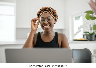 African Woman Smiling, Holding Eye Glasses. Looking At Laptop While Sitting And Working From Home. Wearing Traditional Head Scarf