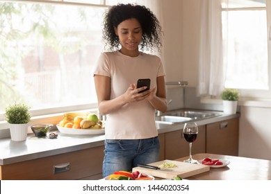 African Woman Standing In Kitchen Distracted From Cooking Preparing Dinner Received Sms Read Message Uses Phone, Searching Recipe, Taking Picture Of Vegetable Dish Social Networks User Blogger Concept