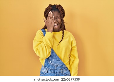 African woman standing over yellow background covering one eye with hand, confident smile on face and surprise emotion.  - Shutterstock ID 2260397539