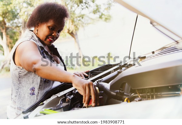 African woman stands by checking\
car battery terminals with car problems. Travel problems waste\
time, pickup truck engine failure. on the road while traveling\
