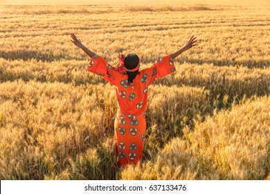 African Woman In Traditional Clothes Standing Arms Raised In A Field Of Barley Or Wheat Crops At Sunset Or Sunrise