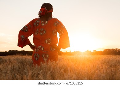 African woman in traditional clothes standing with her hands on her hips in field of barley or wheat crops at sunset or sunrise