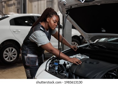 African Woman Under The Hood Of White Car. Woman In Uniform Mechanic Repairing A Car In Car Service. Portrait Of Young Black Female Enjoying Work With Automobile, Vehicle