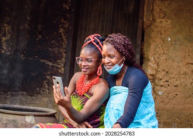 An African woman using glasses with a friend using nose mask happily look into a smart phone outside a village mud house that has a big water calabash