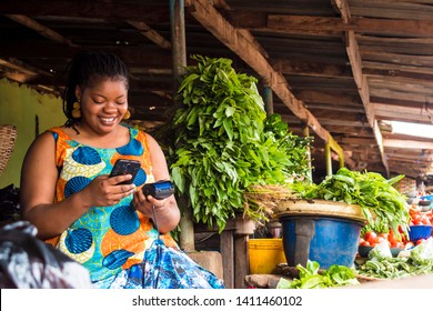 African Woman Using Her Phone And Also Holding A Pos System In A Local Market