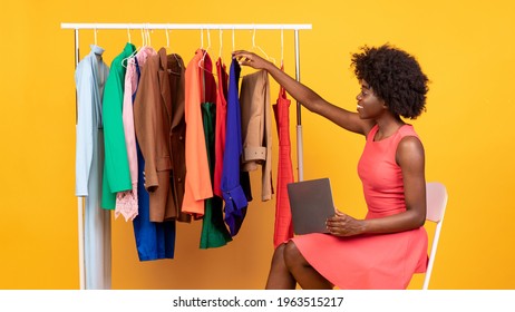 African Woman Using Laptop Writing For Fashion Blog About Shopping Trends Sitting Near Clothing Rack On Yellow Studio Background. Style Assistant Or Clothes Boutique Owner Working In Studio. Panorama