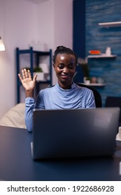 African Woman Waving At Laptop Webcam In The Course Of Video Conference Working Late At Night From Home Office. Black Freelancer Working With Remotely Team Chatting Virtual Online Conference.