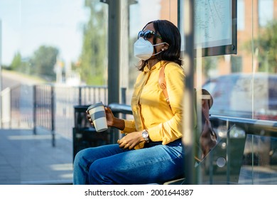 African Woman Wearing Protective Face Mask And Sunglasses In A Bus Station Waiting For The Bus Alone. Black Female Student Sitting On Bench At Public Transportation Stop After Reopening Post Lockdown.