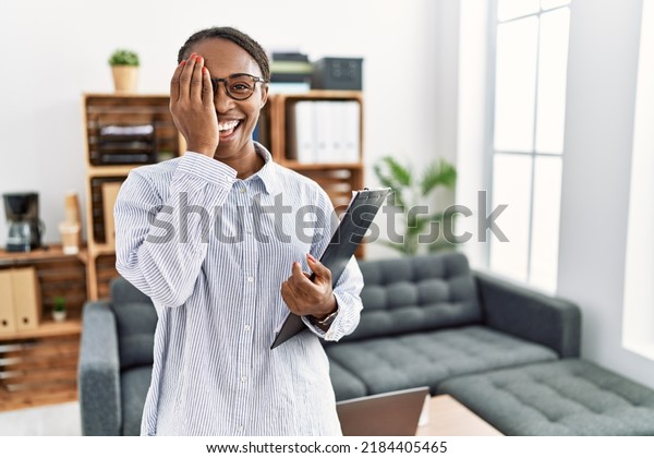 African\
woman working at psychology clinic covering one eye with hand,\
confident smile on face and surprise emotion.\
