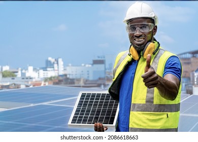 African Worker Holding Solar Cell Panel And Standing Confidence At Rooftop Of Solar Cell Panel In Background. Operation On Site Renewable Energy Construction.