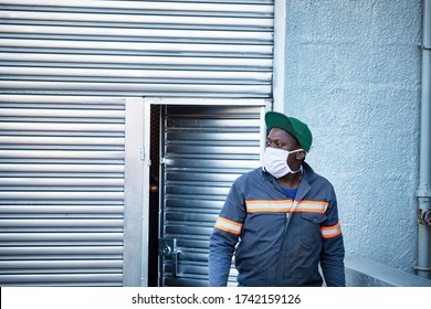 African Worker  Wearing A Mask, Outdoors View, Exiting A Metallic Door At A Warehouse 