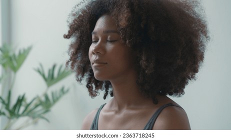 African young adult female engages in a deep meditation or yoga practice at home. With her eyes gently closed, she finds inner peace and calm amidst the demands of daily life.