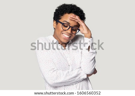 African young bespectacled woman in casual blouse posing indoors she lowers her eyes down and looks embarrassed feels very confused posing isolated on gray studio background, shy modest person concept