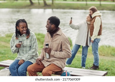 African Young Couple Sitting On Bench And Drinking Tea From Flask With Their Friends Making Selfie In Background At Picnic Outdoors