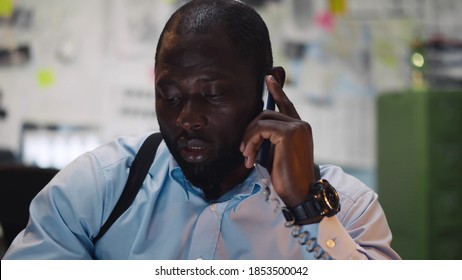 African Young Detective Sitting At Police Department Talking To Colleague On Phone. Portrait Of Black Male Cop Having Phone Call With Witness Investigating Burglary Case In Office