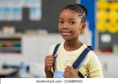 African young girl with blue backpack looking at camera. Pretty and satisfied black schoolgirl with rucksack smiling in class. Portrait of beautiful school girl standing in classroom.