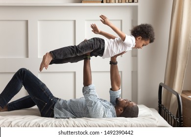 African young loving father lying on bed holding son on raised up hands toddler boy imagines flying in air feels happy not afraid of heights, older younger american brother enjoy time together at home