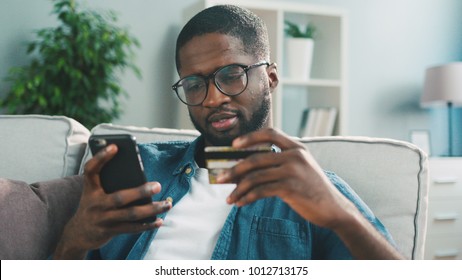 Joven africano en gafas comprando en línea con tarjeta de crédito usando un smartphone en casa. En el interior.
