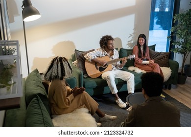 African young man playing guitar sitting on the sofa with his friends singing during their vacations in the house - Powered by Shutterstock