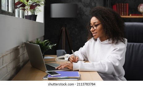 African young teenager e-learning on distance, using laptop, writing homework assignment in copybook, watching webinars online from home. - Shutterstock ID 2113783784