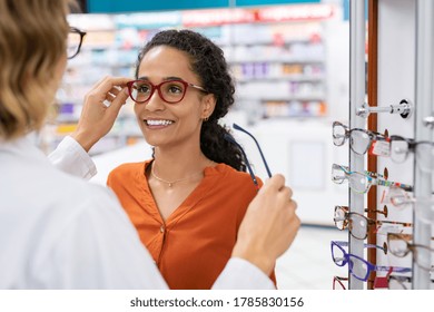 African young woman in optic store choosing new cool-down glasses with optician. Mixed race girl trying new eyeglasses with the help of the pharmacist. Multiethnic woman trying rest spectacle frame.