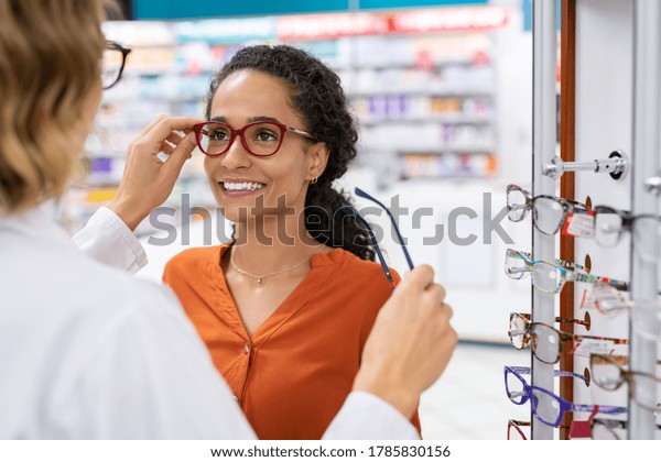 African young woman in optic store choosing new\
cool-down glasses with optician. Mixed race girl trying new\
eyeglasses with the help of the pharmacist. Multiethnic woman\
trying rest spectacle\
frame.