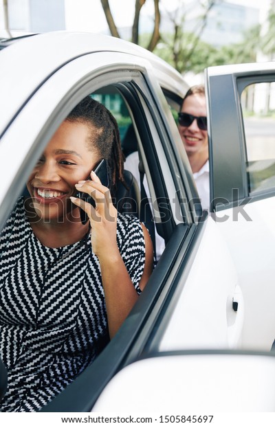 African young woman talking on mobile phone\
and smiling while sitting behind the wheel in car with smiling man\
in the background