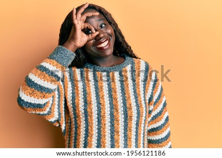 African young woman wearing wool winter sweater smiling happy doing ok sign with hand on eye looking through fingers 