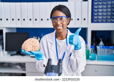 African Young Woman Working At Scientist Laboratory Holding Brain Smiling Happy And Positive, Thumb Up Doing Excellent And Approval Sign 