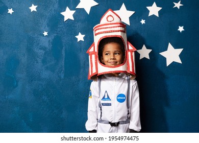 African-american boy playing astronaut with handcrafted rocket helmet on blue studio backdrop with white stars. Portrait of smiling mixed race child dreaming of cosmic space. Childhood and imagination