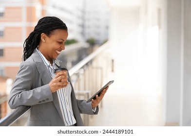 African-American businesswoman standing on a balcony of an office building, taking a break from work, drinking coffee, and using a small tablet computer - Powered by Shutterstock