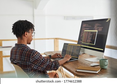 African-American IT developer typing on keyboard with black and orange programming code on computer screen and laptop in contemporary office interior, copy space