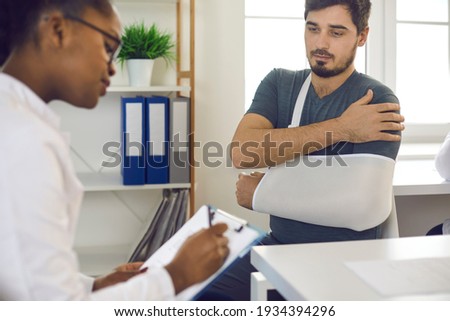 African-American doctor at hospital prescribing treatment and talking to young patient wearing injury support, forearm sling immobilizer, brace, cast, scarf bandage on broken hand, wrist, elbow or arm