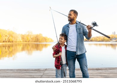 An African-American father and his son stand on a wooden pier by a lake, both holding fishing rods. The father, dressed in a denim jacket and jeans, hugs his son, who is wearing a red plaid shirt and - Powered by Shutterstock