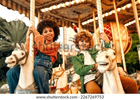 An African-American girl with Afro-curls and a Caucasian curly-haired blonde boy ride a vintage horse carousel in the evening at an amusement park on a weekend.