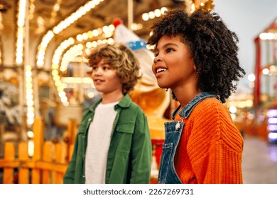 An African-American girl child with afro curls, and a Caucasian boy look admiringly at a carousel of horses at an amusement park or circus on a weekend evening.