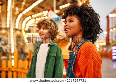 An African-American girl child with afro curls, and a Caucasian boy look admiringly at a carousel of horses at an amusement park or circus on a weekend evening.
