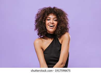 African-American girl with fluffy brunette hairstyle smiles on lilac background. Cool lady in dark top looks into camera on isolated backdrop..