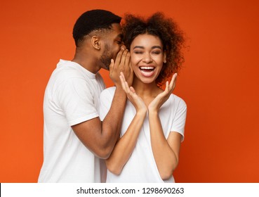 African-american girl laughing, listening how her boyfriend whispering secret or gossips, orange studio background