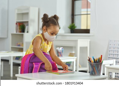 an African-American girl with medical mask on her face puts a copybook on a schooldesk. A child in a yellow dress stands in the classroom and prepares for a lesson - Powered by Shutterstock