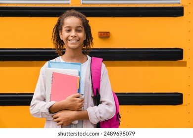 African-american girl teenager pupil student preparing to go to school after summer holidays holding books and notebooks standing next to the school bus. - Powered by Shutterstock