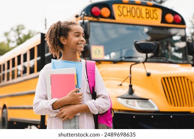 African-american girl teenager pupil student preparing to go to school after summer holidays holding books and notebooks standing next to the school bus. - Powered by Shutterstock