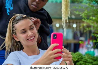 An African-American hairdresser weaves French braids to a smiling girl with a phone in her hands. A professional hairdresser makes a hairstyle for a beautiful young woman - Powered by Shutterstock