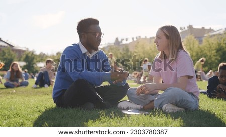 African-American helping girl during outdoor lesson with college students on lawn in park. Portrait of professor and teenage schoolgirl talking sitting on school backyard