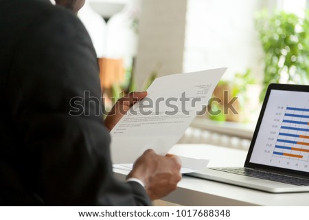 African-american hr manager or employer reading cover letter from job vacancy applicant, black businessman holding business mail checking paper correspondence at workplace, over the shoulder view