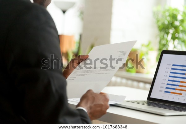 African-american hr manager or employer reading cover letter from job vacancy applicant, black businessman holding business mail checking paper correspondence at workplace, over the shoulder view