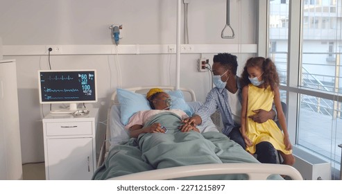 African-American husband and daughter in safety mas visiting sick woman lying in hospital bed. Ill female patient with oxygen mask lying in hospital ward with African-American man and little girl - Powered by Shutterstock